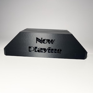 May include: A black, angled stand with the words "Now Playing" cut out of the front. The stand is designed to hold a phone or tablet. The stand is made of a matte black material.