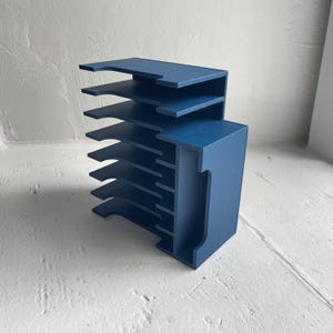May include: A blue wooden desk organizer with multiple shelves and a vertical section. The organizer has a modern design and is painted in a solid, dark blue color. The shelves are spaced evenly, creating a tiered structure.