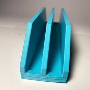 May include: A turquoise 3D-printed desk organizer with three vertical slots. The organizer is rectangular with a textured surface and is designed to hold papers, mail, or other office supplies. The object is made of plastic.