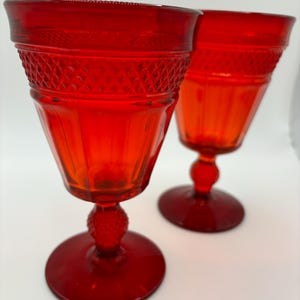 May include: Two ruby red goblets with a textured diamond pattern around the rim and a round base. The stem has a textured, spherical design. The glasses are set against a white background.