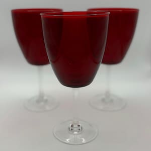 May include: Three ruby red wine glasses with clear stems and bases. The glasses are arranged in a slightly offset triangular formation against a white background. The glasses have a classic, elegant design.