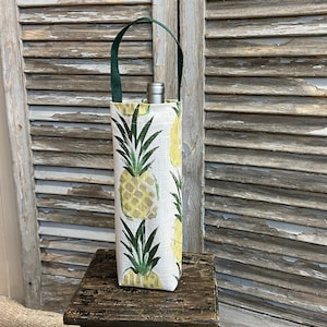 May include: A white wine bottle carrier with a green handle and a pineapple print. The carrier is sitting on a wooden stool.
