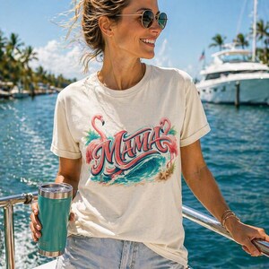 Puede incluir: Camiseta color crema con un diseño tropical que presenta la palabra "Mama" en escritura rosa, flanqueada por flamencos y olas del océano. La persona sostiene un vaso de color turquesa. El fondo muestra un barco y el océano.