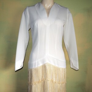 May include: A white dress with long sleeves and a V-neckline. The dress has a fitted bodice and a skirt with a fringe trim.