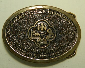 May include: A gold-toned metal belt buckle with the text "AMAX COAL COMPANY WESTERN DIVISION SIGNIFICANT CONTRIBUTION TO SAFETY 1982". The buckle features a logo with a stylized image of a building, gears, and people.