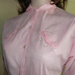 May include: A pink button-down shirt with a peter pan collar and pearl-like buttons. The shirt has a single pocket on the left side.
