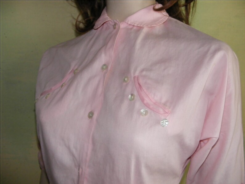 May include: A pink button-down shirt with a peter pan collar and pearl-like buttons. The shirt has a single pocket on the left side.
