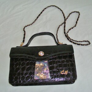May include: A black crocodile-embossed leather handbag with a gold chain strap and a gold clasp. The bag has a small iridescent patch on the front and the letters "C#" in gold.