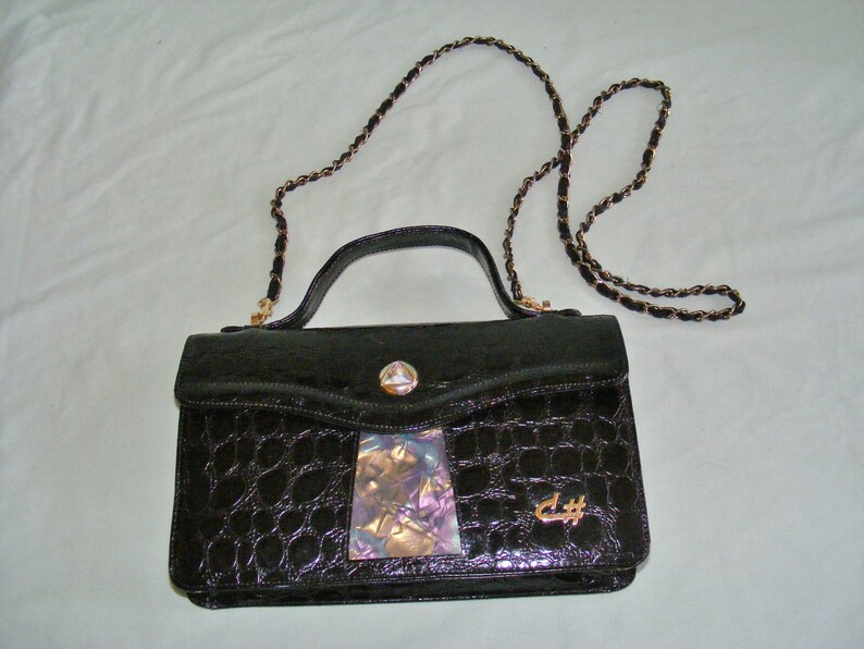 May include: A black crocodile-embossed leather handbag with a gold chain strap and a gold clasp. The bag has a small iridescent patch on the front and the letters "C#" in gold.