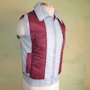 May include: A grey and burgundy quilted vest with a zip closure and two pockets on the front. The vest has a stand-up collar.