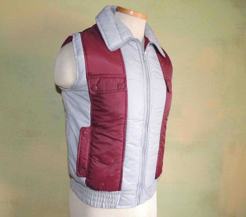 May include: A grey and burgundy quilted vest with a zip closure and two pockets on the front. The vest has a stand-up collar.