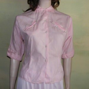 May include: A pink button-down shirt with a collar and short sleeves. The shirt has pearl-like buttons and a slightly gathered look at the shoulders.