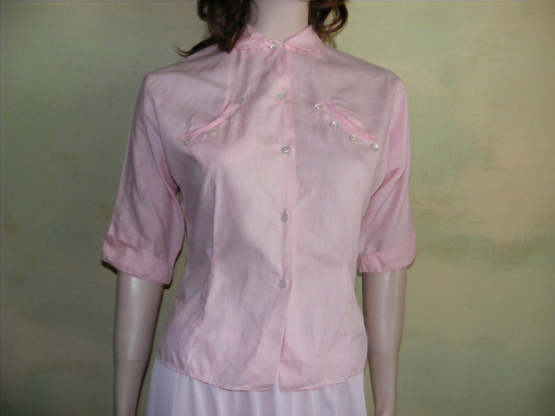 May include: A pink button-down shirt with a collar and short sleeves. The shirt has pearl-like buttons and a slightly gathered look at the shoulders.
