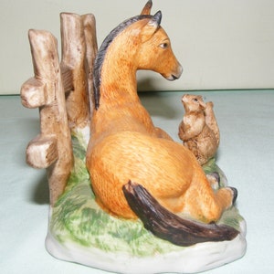 May include: A ceramic figurine of a brown horse lying down next to a wooden fence with two brown squirrels sitting on the fence.
