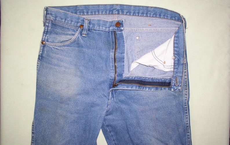 May include: A pair of blue denim jeans with a zipper fly and a button closure. The zipper is open and the fly is turned back to reveal the inside of the jeans.