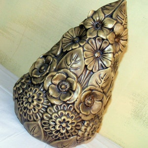 Puede incluir: Un jarrón de cerámica con forma de cono y superficie texturizada. El jarrón está decorado con flores y hojas en relieve en tonos marrones y dorados.