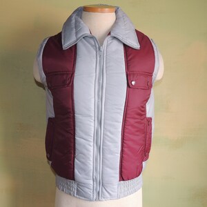 May include: A grey and burgundy quilted vest with a zip closure and two pockets. The vest has a stand-up collar.