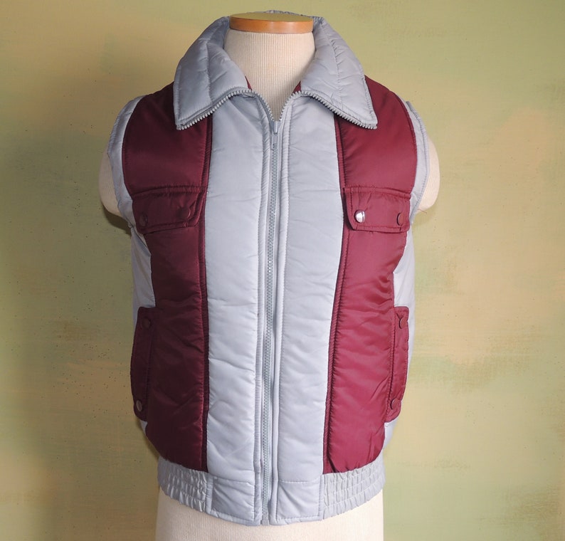May include: A grey and burgundy quilted vest with a zip closure and two pockets. The vest has a stand-up collar.