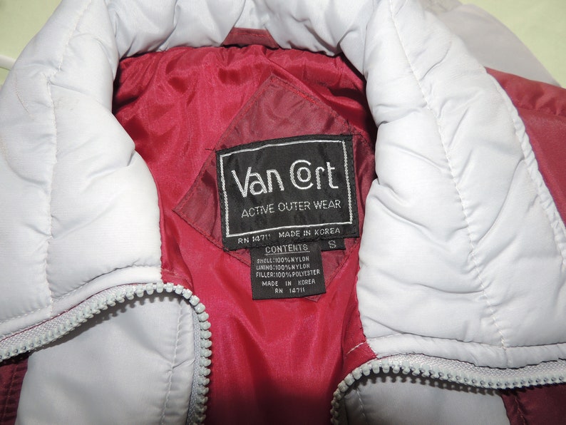 May include: A red and white puffer jacket with a black label that reads "Van @rt Active Outer Wear" and "RN 14711 MADE IN KOREA". The label also lists the contents as "SHELL: 100% NYLON, LINING: 100% NYLON, FILLER: 100% POLYESTER, MADE IN KOREA, RN 14711".