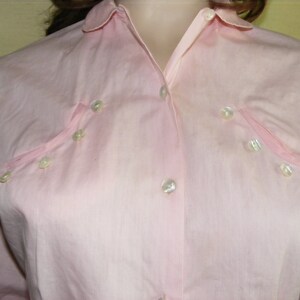 May include: A pale pink button-down shirt with a collar and long sleeves. The shirt has a row of pearl-like buttons down the front and a few more buttons on the chest.