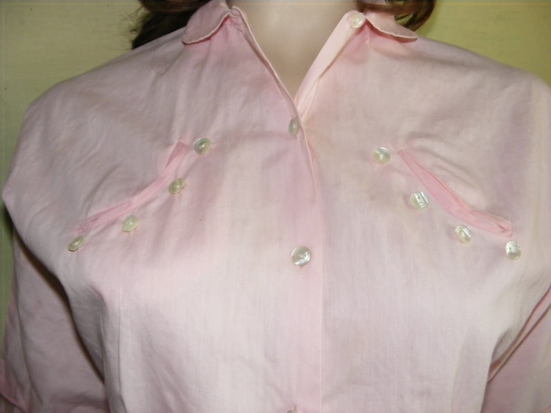 May include: A pale pink button-down shirt with a collar and long sleeves. The shirt has a row of pearl-like buttons down the front and a few more buttons on the chest.