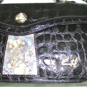 May include: Black leather handbag with a crocodile-embossed pattern. The bag has a flap closure with a silver-toned clasp and a decorative silver-toned musical note charm. The bag also features a clear plastic window on the front.