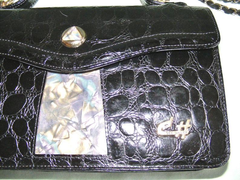 May include: Black leather handbag with a crocodile-embossed pattern. The bag has a flap closure with a silver-toned clasp and a decorative silver-toned musical note charm. The bag also features a clear plastic window on the front.