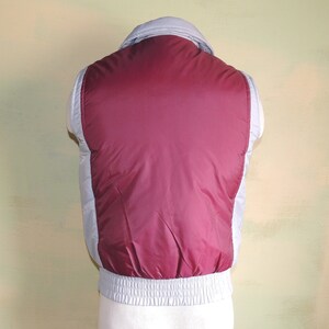 May include: A sleeveless puffer vest with a burgundy front and a light grey back. The vest has a zip closure and a ribbed waistband.