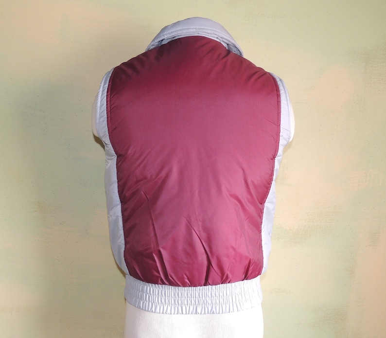 May include: A sleeveless puffer vest with a burgundy front and a light grey back. The vest has a zip closure and a ribbed waistband.