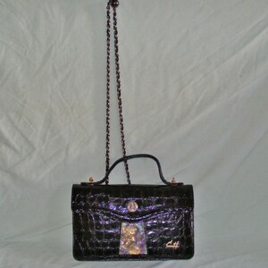 May include: A black crocodile-embossed handbag with a gold chain strap and a gold clasp. The bag has a small, decorative patch on the front with a floral design.