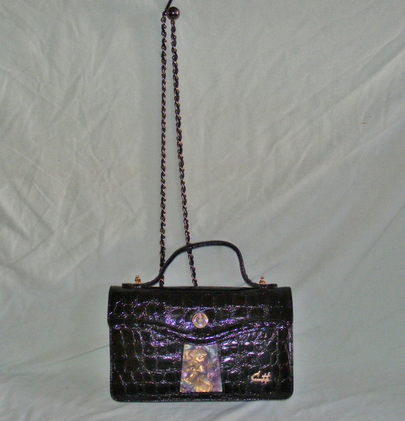 May include: A black crocodile-embossed handbag with a gold chain strap and a gold clasp. The bag has a small, decorative patch on the front with a floral design.