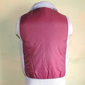 May include: A sleeveless puffer vest with a burgundy front and a light grey back. The vest has a zip closure and a stand-up collar.