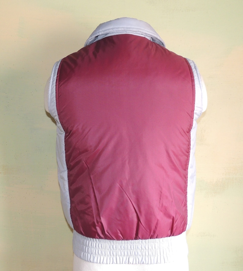 May include: A sleeveless puffer vest with a burgundy front and a light grey back. The vest has a zip closure and a stand-up collar.
