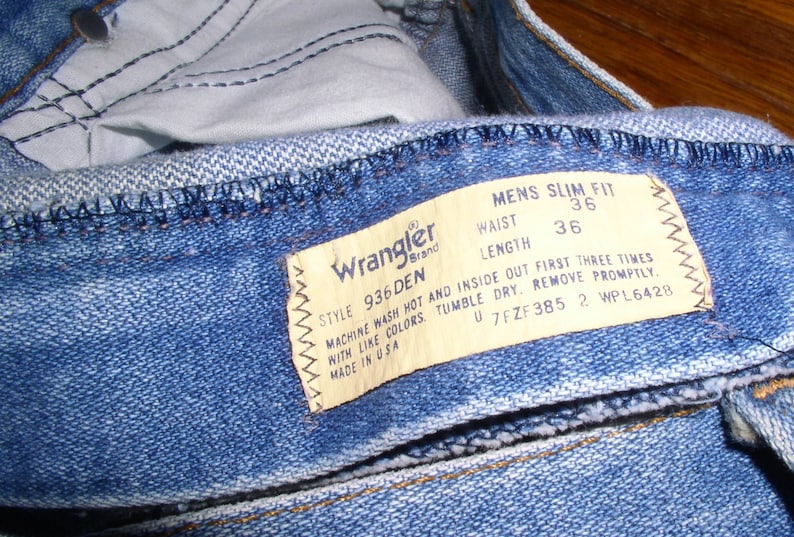 May include: A blue denim jean with a Wrangler brand label. The label states "Style 936 Den" and "Mens Slim Fit". The waist size is 36 inches and the length is 36 inches. The label also includes instructions for washing and drying the jeans.