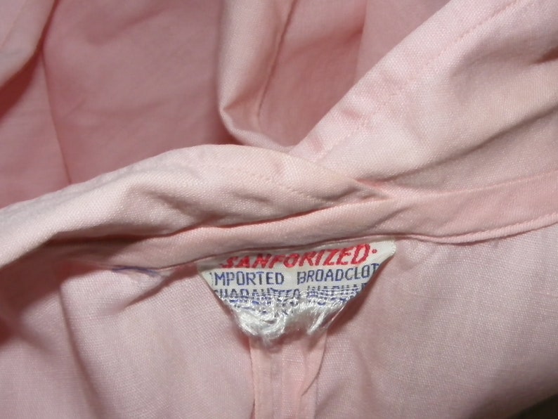 May include: A close-up of a pink fabric label with the text "Sanforized. Imported Broadcloth" in blue.
