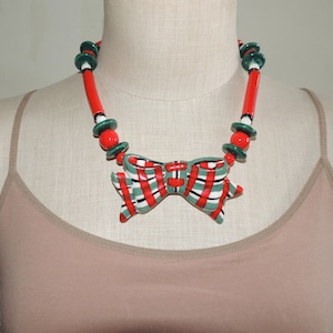 May include: A red, green, and white ceramic bow necklace. The bow is the centerpiece of the necklace and is surrounded by red and green beads.