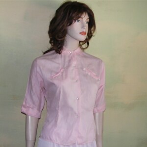May include: A pink button-up shirt with a mandarin collar and pearl-like embellishments. The shirt has three-quarter sleeves and a fitted silhouette.