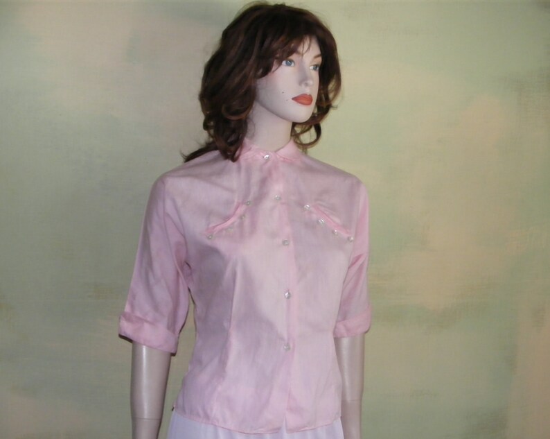 May include: A pink button-up shirt with a mandarin collar and pearl-like embellishments. The shirt has three-quarter sleeves and a fitted silhouette.
