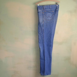 May include: A pair of blue denim jeans hanging on a wooden hanger against a light green wall.