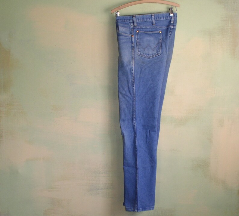 May include: A pair of blue denim jeans hanging on a wooden hanger against a light green wall.