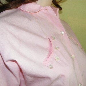 May include: A pink button-down shirt with a collar and a pocket on the left side. The shirt has pearl-like buttons.