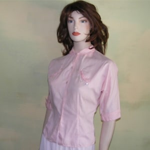 May include: A pink button-up shirt with short sleeves and a collar. The shirt has two pockets on the front with decorative stitching and pearl-like embellishments.