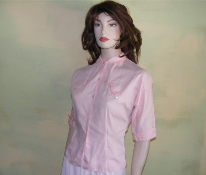May include: A pink button-up shirt with short sleeves and a collar. The shirt has two pockets on the front with decorative stitching and pearl-like embellishments.