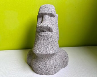 Könnte beinhalten: Eine graue Steinskulptur eines Moai-Kopfes, eine traditionelle Statue von der Osterinsel. Die Skulptur ist detailliert mit einem geschnitzten Gesicht und einer strukturierten Oberfläche.
