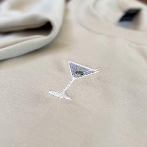 Puede incluir: Sudadera de cuello redondo beige claro con un pequeño diseño bordado de copa de Martini. La copa de Martini presenta un líquido azul, una aceituna verde y un tallo blanco.
