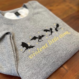 May include: A gray sweatshirt with a crew neck, featuring a design of black dragons and the text "violent little thing" in yellow. The sweatshirt is folded, showing the design on the front.