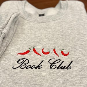 May include: A light gray sweatshirt with embroidered red chili peppers and the words "Book Club" in black script. The sweatshirt is folded, showing the sleeve and neckline.
