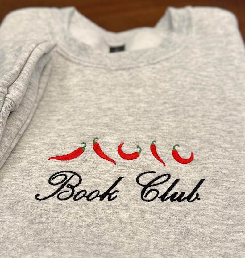 May include: A light gray sweatshirt with embroidered red chili peppers and the words "Book Club" in black script. The sweatshirt is folded, showing the sleeve and neckline.