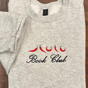 May include: A light gray sweatshirt with embroidered red chili peppers and the words "Book Club" in black script. The sweatshirt is folded neatly on a wooden surface.