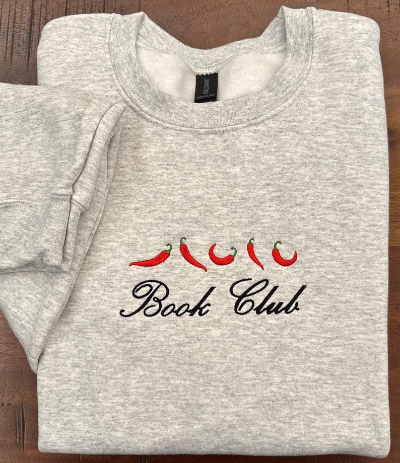 May include: A light gray sweatshirt with embroidered red chili peppers and the words "Book Club" in black script. The sweatshirt is folded neatly on a wooden surface.